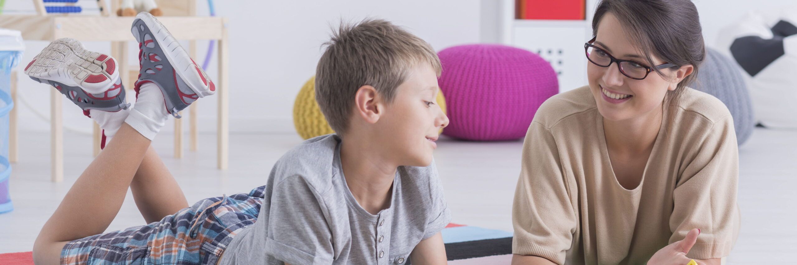 A young boy and a woman smiling at each other while sitting on the floor in a colorful psychiatric services playroom, with toys and a bean bag in the background.