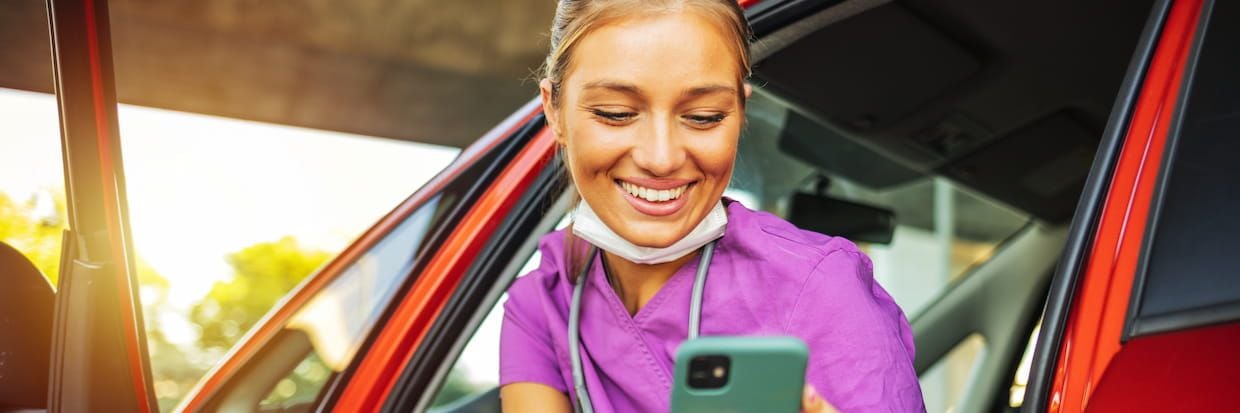 A smiling registered nurse in a purple uniform looks at her smartphone inside a car, with her face mask pulled down to her chin, sunlight illuminating her face.