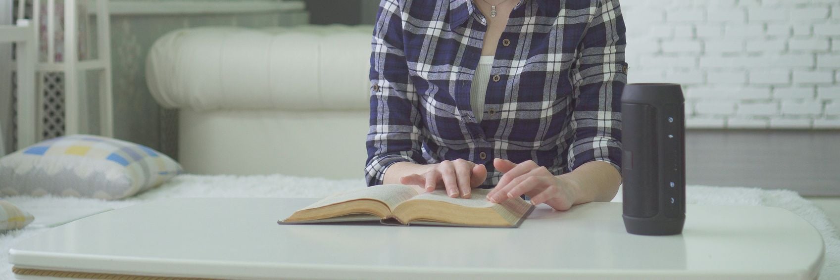 A person in a plaid shirt sits at a white table, reading a book with a black smart speaker beside them in a cozy room.