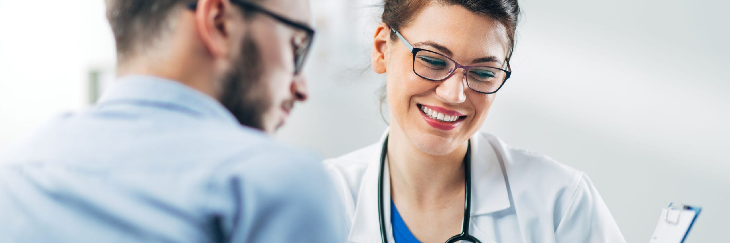 A female doctor with glasses and a stethoscope smiling at a male patient, both looking at a document the doctor is holding during a consultation in a bright, modern office.