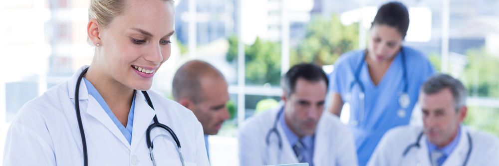A female doctor in a white coat smiles as she reviews her tablet, with three colleagues discussing in the background. the setting is a bright medical office.
