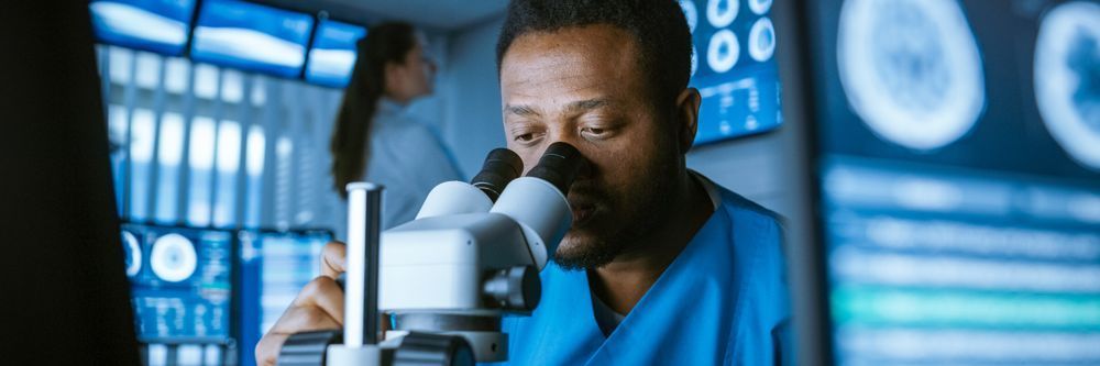 A focused male scientist in a blue lab coat examines samples through a microscope in a high-tech lab, with a female colleague working in the background.
