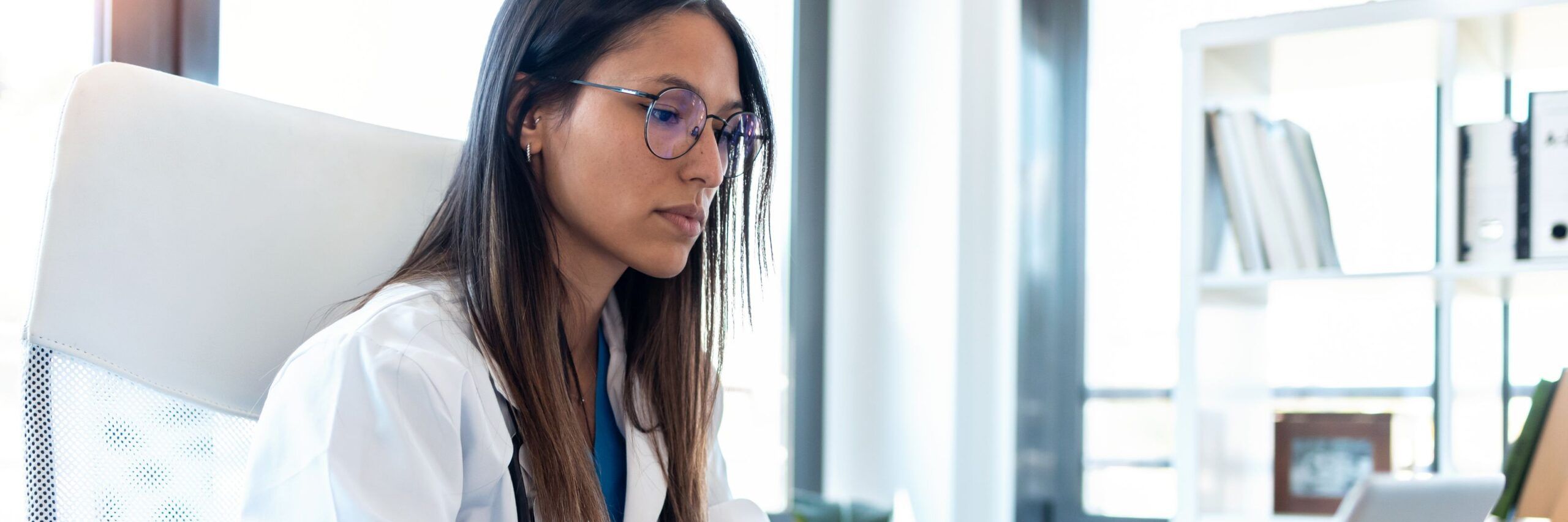 A focused woman in a white blouse and glasses sitting at a desk, reading a document about scholarships in a bright office environment.