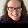 A smiling woman with long, wavy brown hair wearing glasses and a purple jacket over a plaid shirt. She appears joyful and is looking directly at the camera.