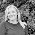 Black and white portrait of a smiling woman with blonde hair, wearing earrings and a dark sweater, standing in front of lush foliage.